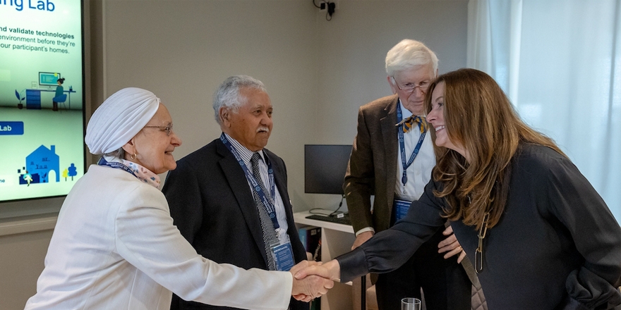 A diverse group of four people, including an older woman wearing a white head covering, two senior men, and a former MP Gillian Keegan, are shaking hands and engaging in conversation in front of a display about a "Living Lab" that tests technologies for home environments. 