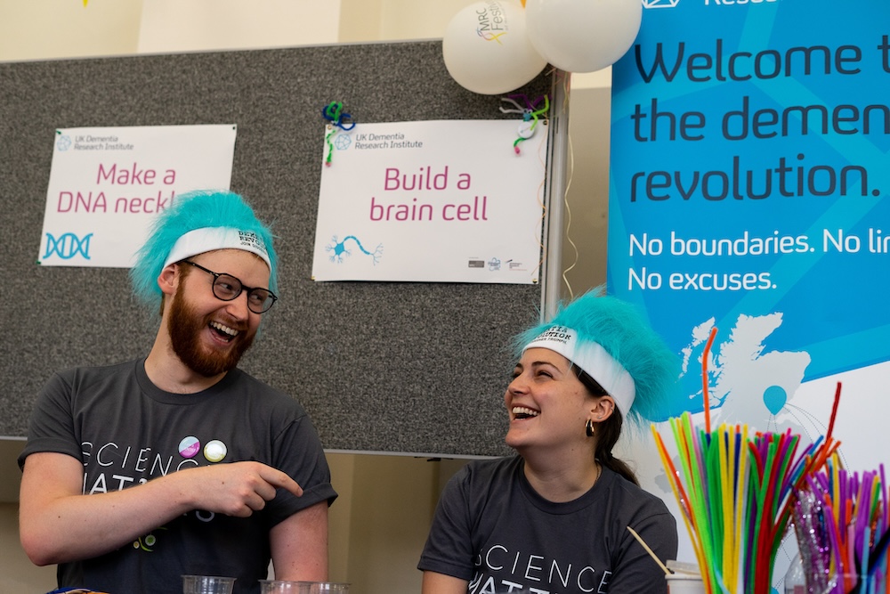 Two enthusiastic volunteers wearing matching "Science" t-shirts and blue-and-white hats are laughing together at an interactive dementia research event, with activity signs and colourful crafting materials visible in the background.