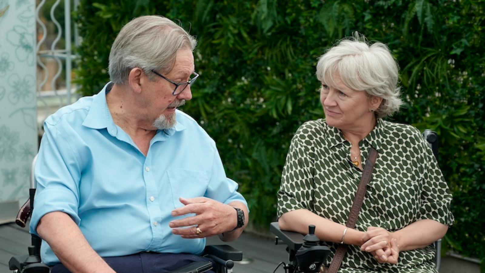 A man and a woman in wheelchairs, engage in conversation outdoors against a backdrop of green foliage, with the man in a blue shirt gesturing as he speaks to the woman in a patterned green dress.