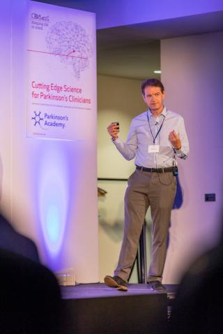 Male presenting in front of a banner reading 'Cutting edge science for Parkinson's clinicians'