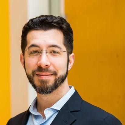 Male with dark hair and a dark beard, and glasses against an orange background