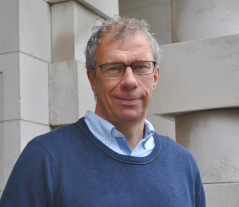 Male with grey hair and glasses, stood against a building wall