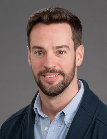 Young man with dark hair and dark facial hair, wearing a light blue shirt