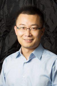 Male with dark hair, glasses and blue shirt against a dark background