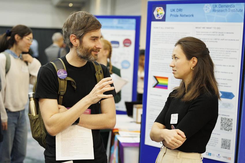 Two people stand in conversation in front of a poster that reads 'UK DRI pride network'
