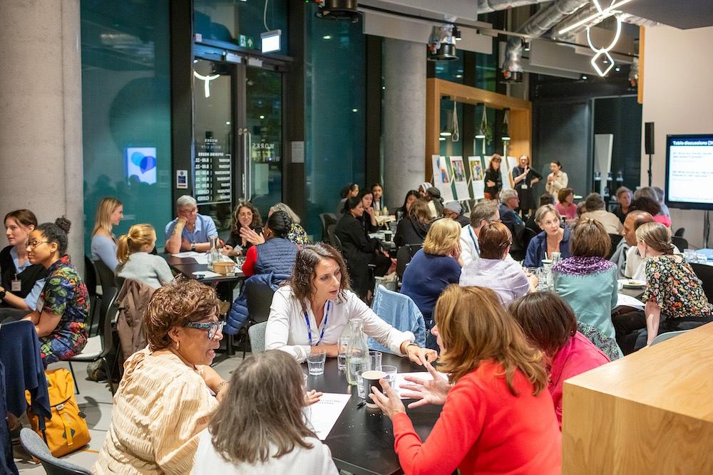 A crowded room filled with diverse groups of people seated at tables, engaged in discussions during what appears to be a conference or networking event in a modern, well-lit venue.