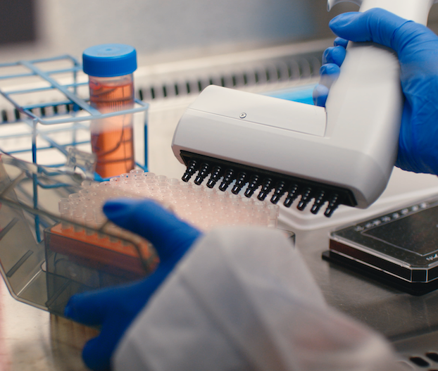 A close-up view of a laboratory workstation showing gloved hands operating a multi-channel pipette, with test tubes, a sample rack, and other scientific equipment visible in the background.