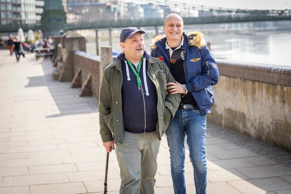 Two men, one with a walking stick, walk arm-in-arm along a riverside promenade on a sunny day, with a bridge visible in the background.