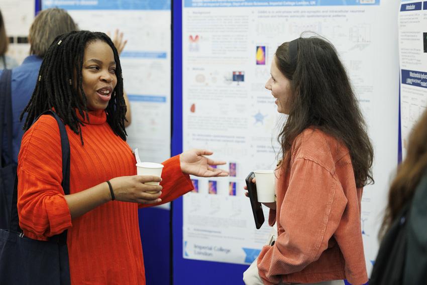 Two women engaged in conversation in front of scientific posters at a conference, both holding cups of coffee, with one wearing a red sweater and the other in an orange jacket.