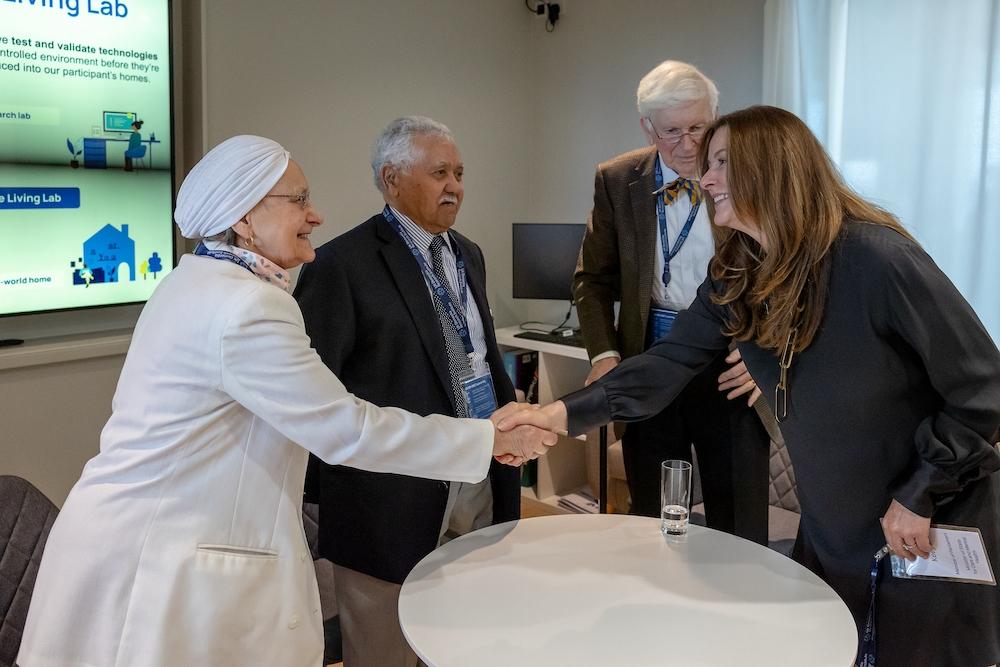 A diverse group of four people, including an older woman in a white head covering, two senior men, and former MP Gillian Keegan, are greeting each other in a room with a "Living Lab" display that showcases technologies for home environments.