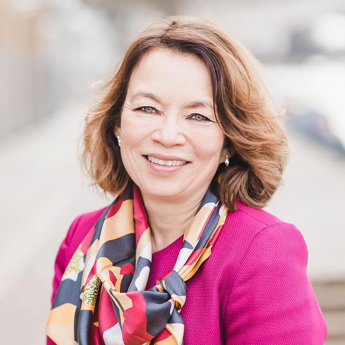 Smiling female with wavy brown hair, wearing a pink blouse and colourful scarf